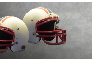  Two vintage white American football helmets with red facemasks and orange and blue striping against a grey background.