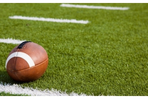 An American football with black laces rests next to the yard line and hashmarks of an American football field.