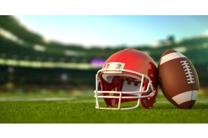 A red American football helmet with a white facemask sitting on grass field in a stadium with a football learning against it.