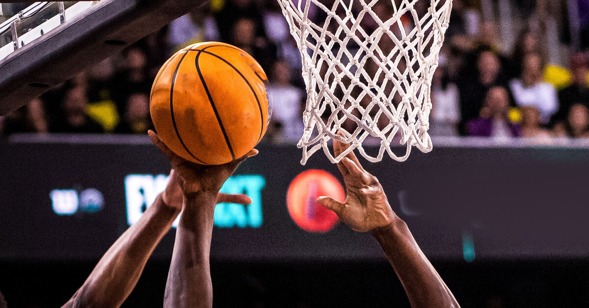 A close-up of the basket, net, and a basketball during a game. Multiple hands reach up for the ball coming off the rim.