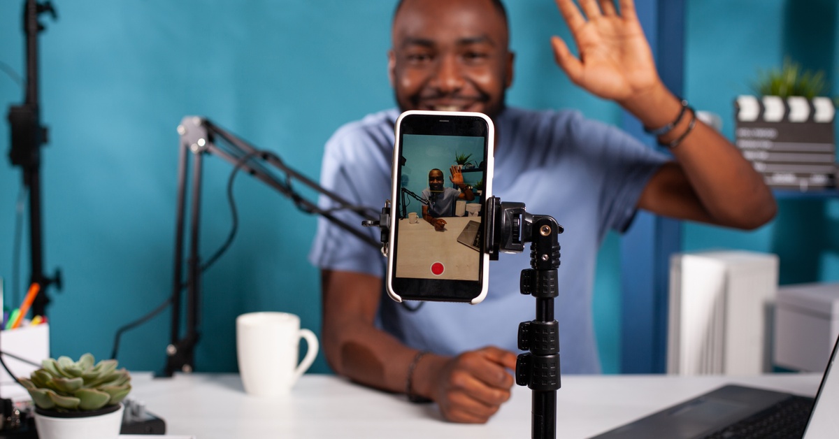A close-up of a smart phone on a stand filming someone at their desk with a microphone waving at the camera.
