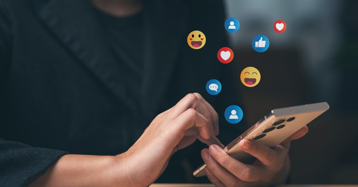 A close-up of a person in black clothes holding and looking at a smartphone with various emojis floating above it.