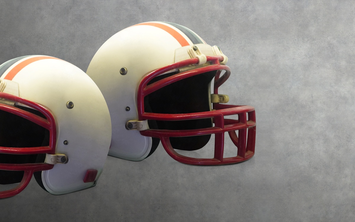  Two vintage white American football helmets with red facemasks and orange and blue striping against a grey background.