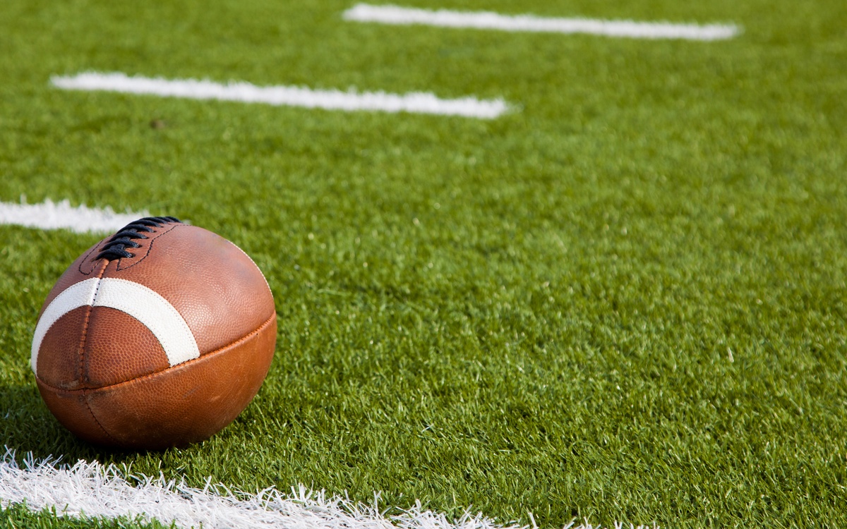An American football with black laces rests next to the yard line and hashmarks of an American football field.