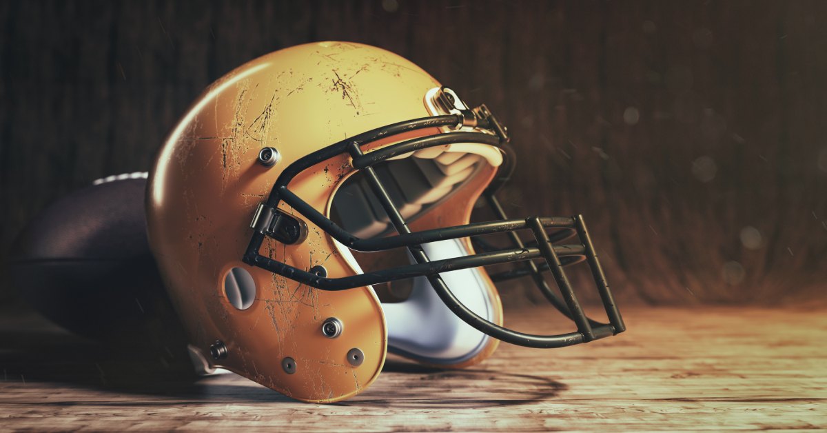 A close-up of a yellow classic American football helmet with scratches on a wooden table in front of a football.