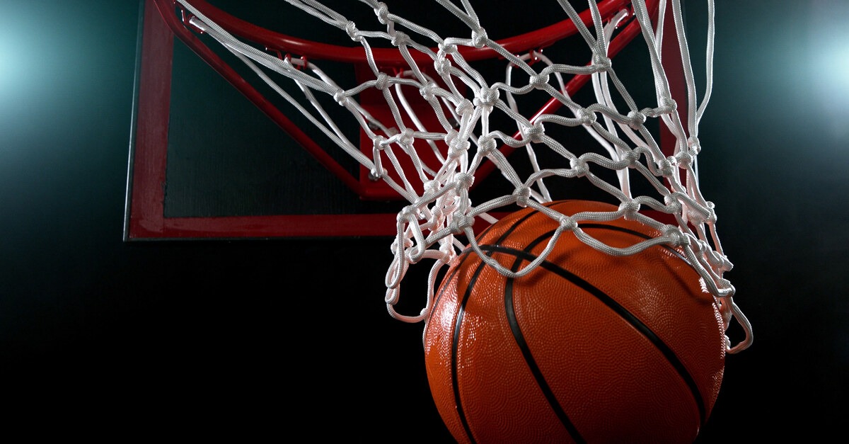 A basketball passing through the white net of a red-orange basketball hoop against a black background.