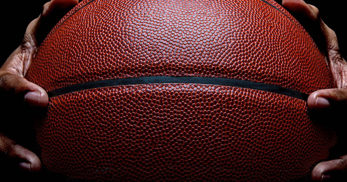 Two large hands gripping an orange basketball by its sides in the dark and against a black background.