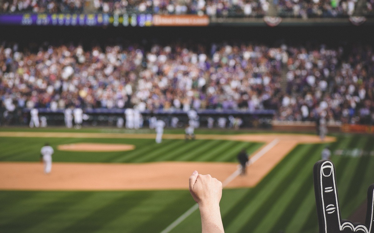 A baseball stadium is full of cheering fans. The focus is on one fan's raised fist, and the field is blurry in the background.